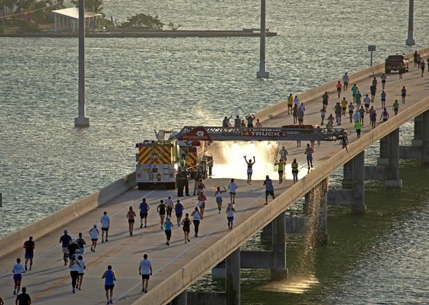 Fotografía cedida hoy por la Oficina de Noticias de los Cayos de Florida donde se muestra a unos corredores mientras pasan por debajo de la escalera de un camión de bomberos adaptada para rociar agua durante su participación en la carrera del Puente de las Siete Millas en los Cayos de Florida (EE.UU).