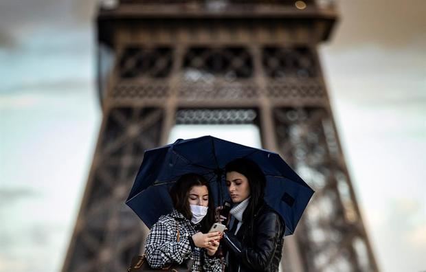 Dos jóvenes cobijadas bajo un paraguas miran su teléfono móvil frente a la Torre Eiffel en París, Francia.