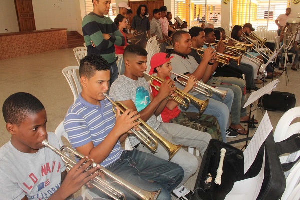  En primer plano, el maestro Samuel Ureña Gonzáles acompaña a los jóvenes en su práctica de trompeta, donde se evidencia la concentración y la entrega de cada uno. En el fondo, un grupo de trombones, tuba y percusión en la misma armonía de los ejercicios.