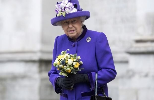 Abrigo y sombrero púrpura intenso con flores textiles blancas y violetas, broche de gema violeta y guantes negros para un evento oficial.