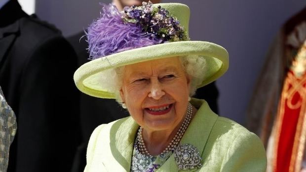 Abrigo verde claro y sombrero decorado con plumas y flores violetas, más collar triple de perlas y broche plateado.