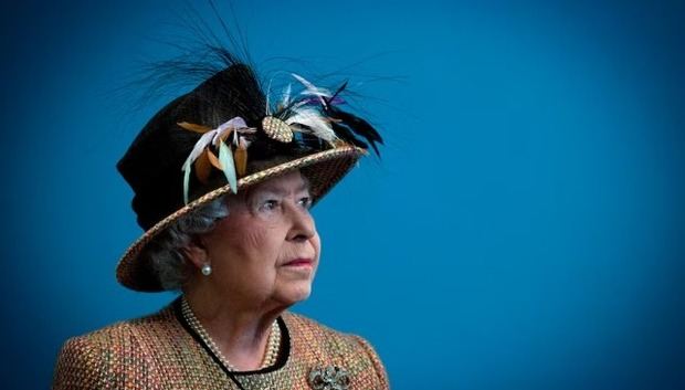 Abrigo jaspeado en tonos tierra y sombrero negro recto, decorado con plumas multicolores y malla central, acompañado de perlas y broche plateado.