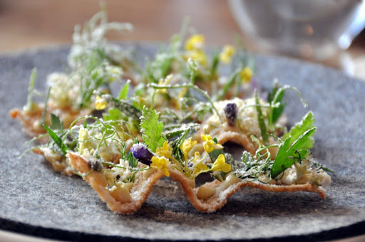 Aperitivo con unas finas tostadas de huevas de rape, acompañadas con flores violetas y polvo de vinagre, lo cual le da la apariencia de un jardín recién tocado por el rocío.